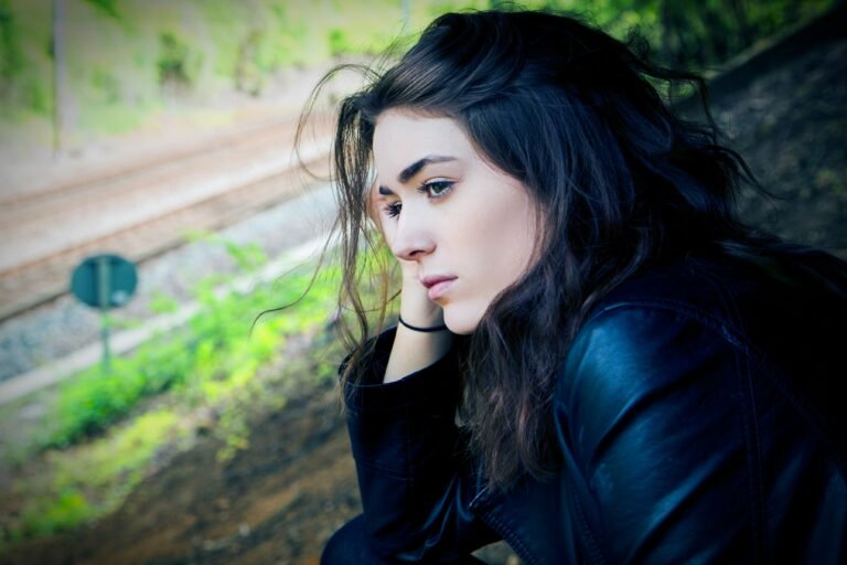 Woman with dark hair in a dark jacket resting her chin on her hand with a thoughtful expression, greenery behind her