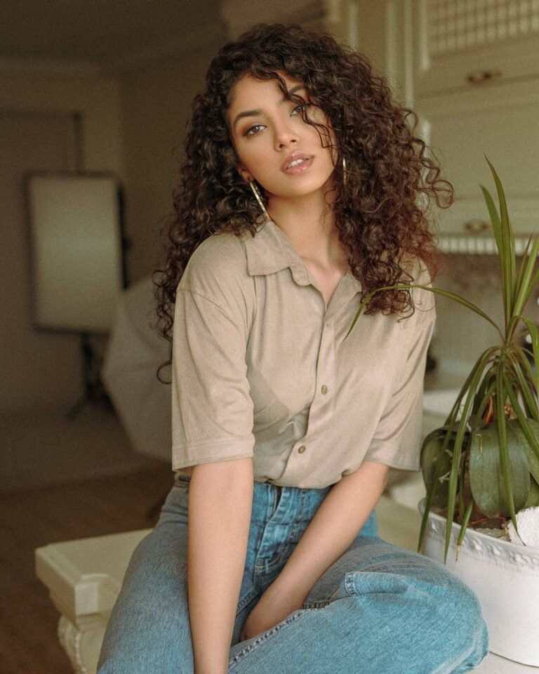 Woman with long curly brown hair in a beige button-up shirt, standing beside a houseplant
