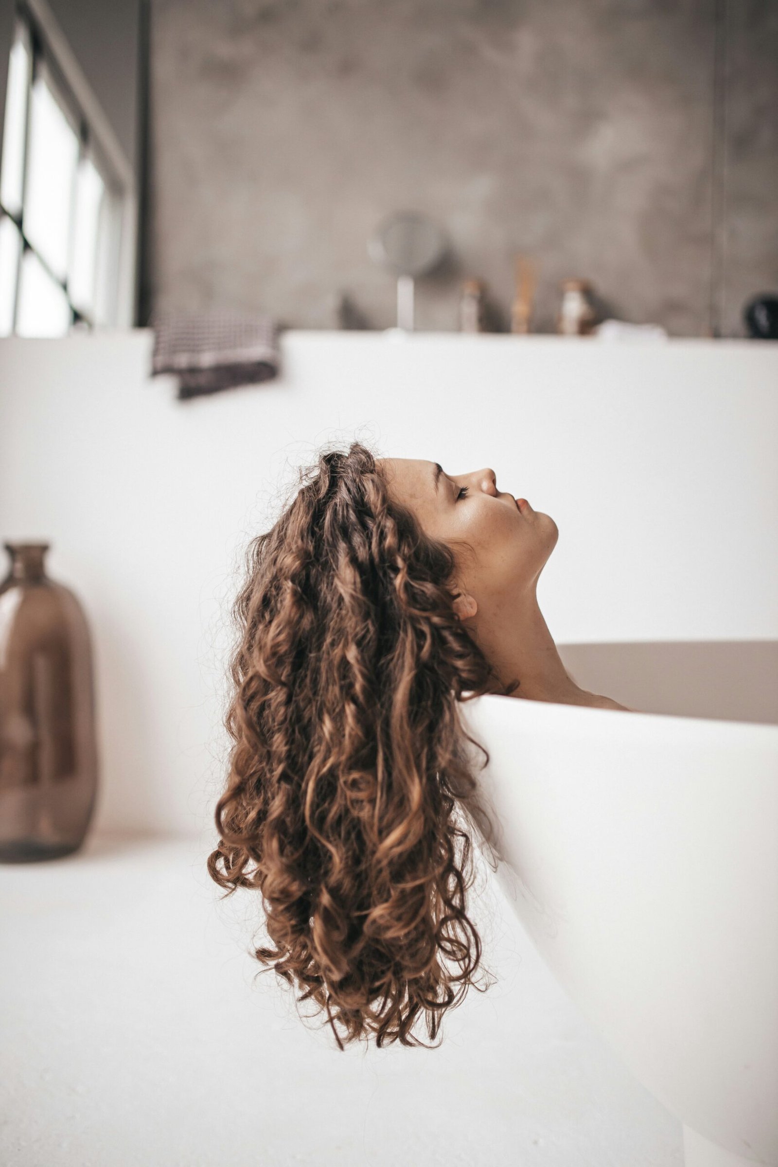 Woman with curly hair leaning her head back in a bathtub, hair flowing over the edge
